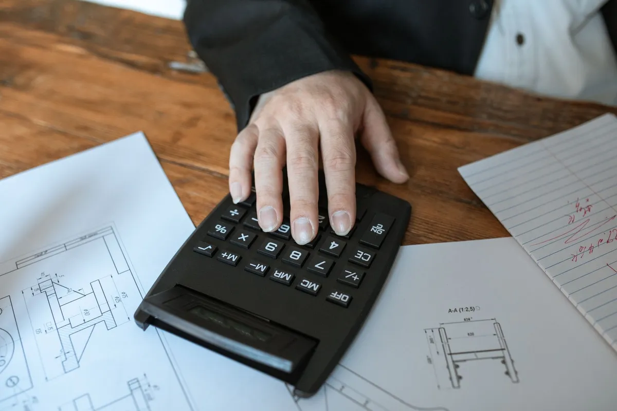 A hand using a calculator on blueprints, symbolizing precision in financial analysis for currency valuation.